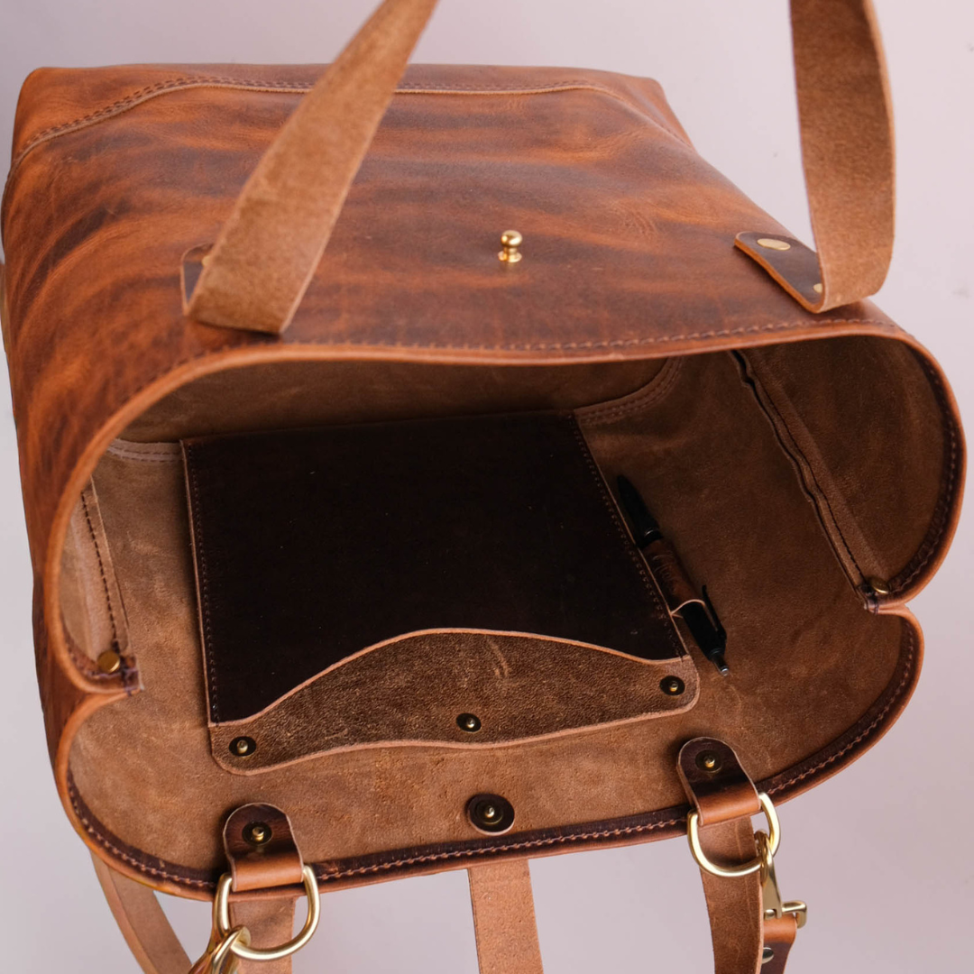 A close-up of an open brown leather bag showing its interior, including a dark insert pocket, a pen loop, brass-tone hardware, and a strap hanging down against a plain light background.