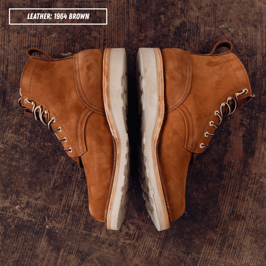 A pair of brown suede lace-up ankle boots shown from above on a dark wood surface, with a text label in the top-left corner reading 'LEATHER: 1964 BROWN'.