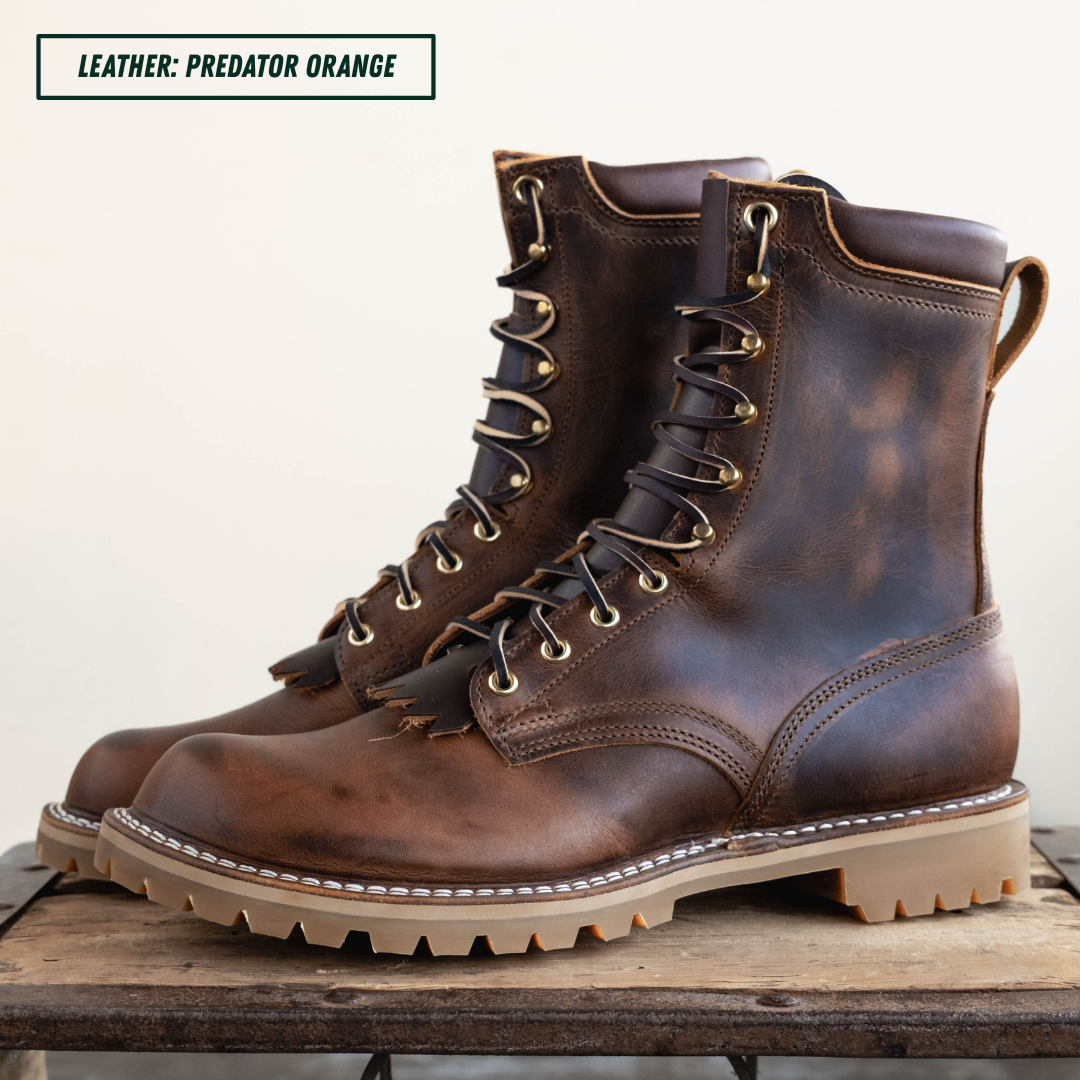 A pair of distressed brown leather lace-up boots with tan soles and brass eyelets, displayed on a rustic wooden surface against a light background. A text label in the top left reads 'LEATHER: PREDATOR ORANGE'.