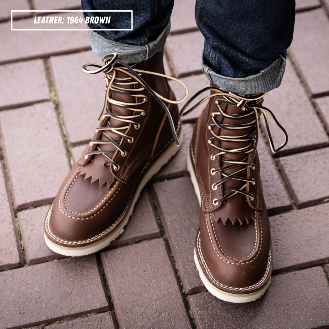 Model wearing Moctoepro boots on brick sidewalk. Label on top left corner that reads "LEATHER: 1964 BROWN."