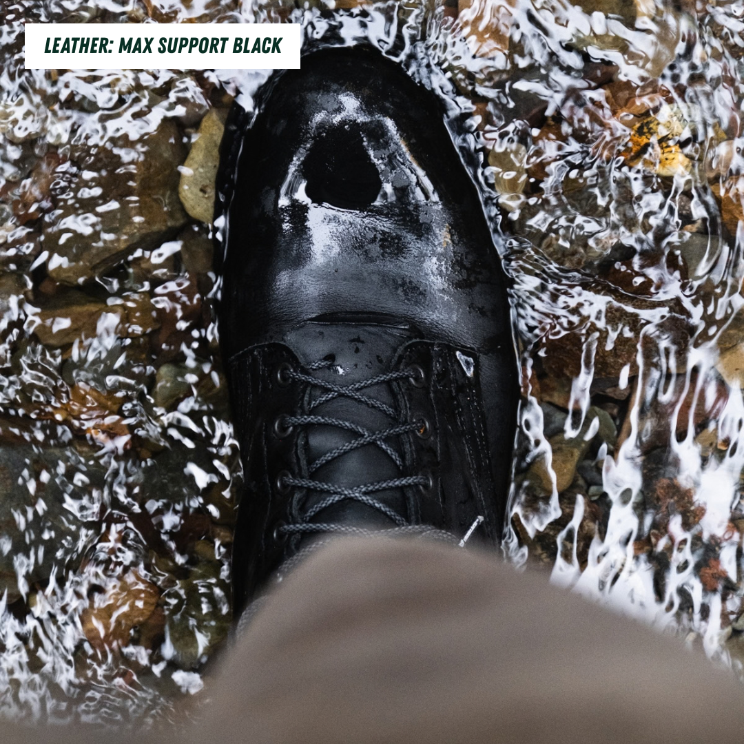 A model wearing a black boot standing in a creek. Top left reads 'LEATHER: MAX SUPPORT BLACK'