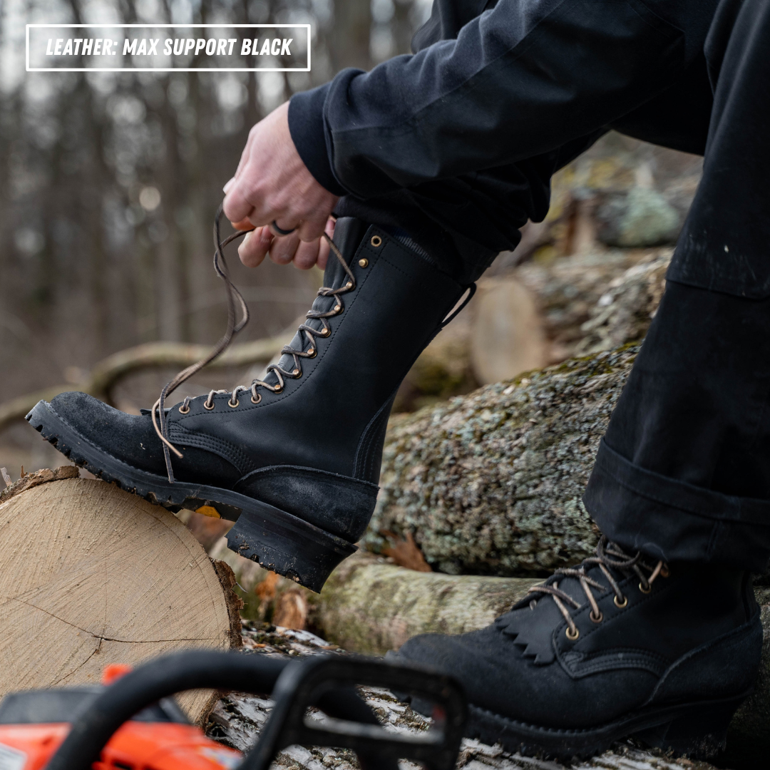A person outdoors by a log wearing a black lace-up work boot and tying the laces. The boot is shown in use on a forest floor with tree trunks and branches in the background.
