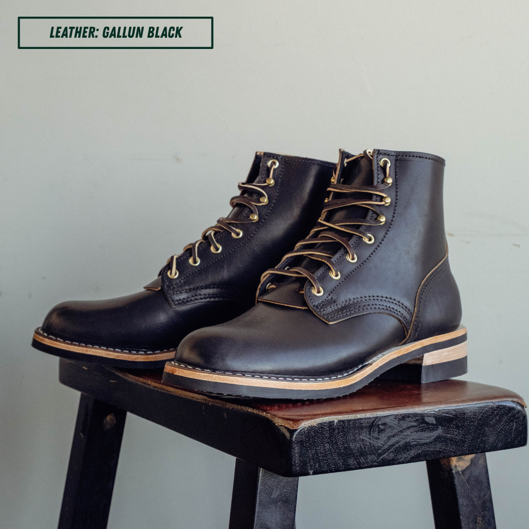 A pair of dark brown leather lace-up boots with tan laces and gold-toned eyelets, displayed on a rustic wooden stool against a light gray wall. A text label in the top left reads 'LEATHER: GALLUN BLACK'.