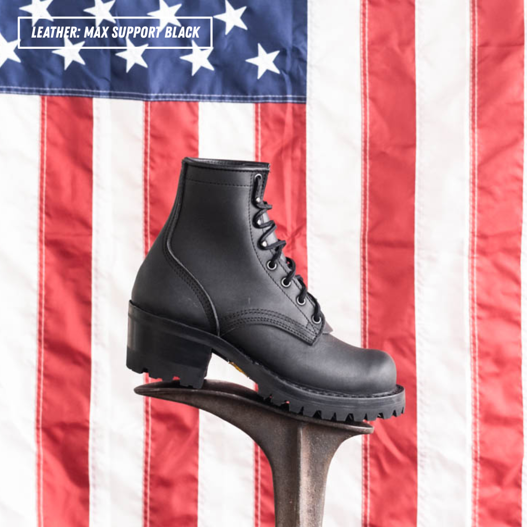 A black leather lace-up ankle boot displayed on a metal stand in front of an American flag backdrop. A text overlay in the top left reads 'LEATHER: MAX SUPPORT BLACK'.