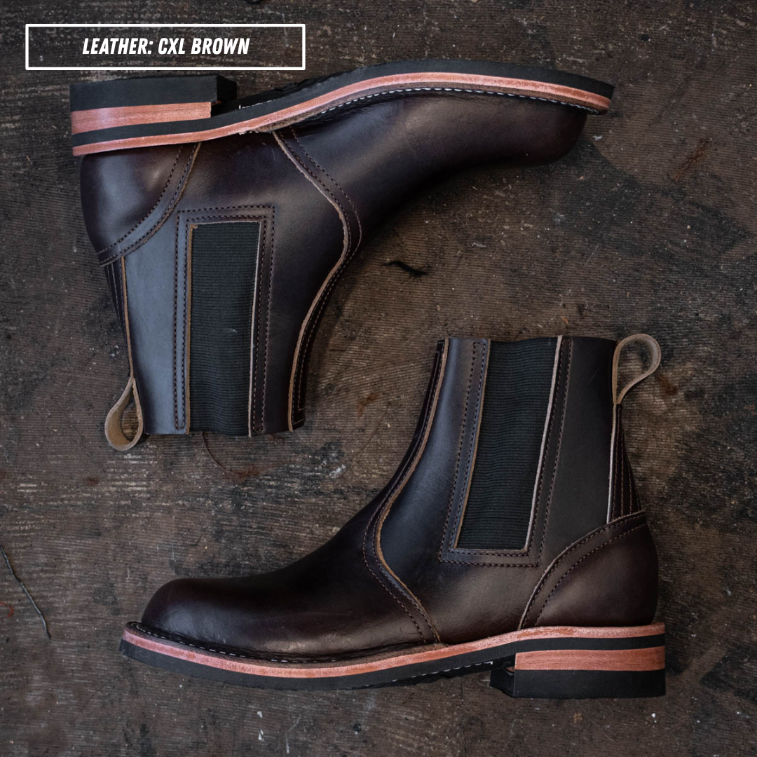 A pair of dark brown leather Chelsea boots with black elastic side panels and a tan welt, arranged on a dark rustic wooden surface. A text overlay in the top left reads 'LEATHER: CXL BROWN'.