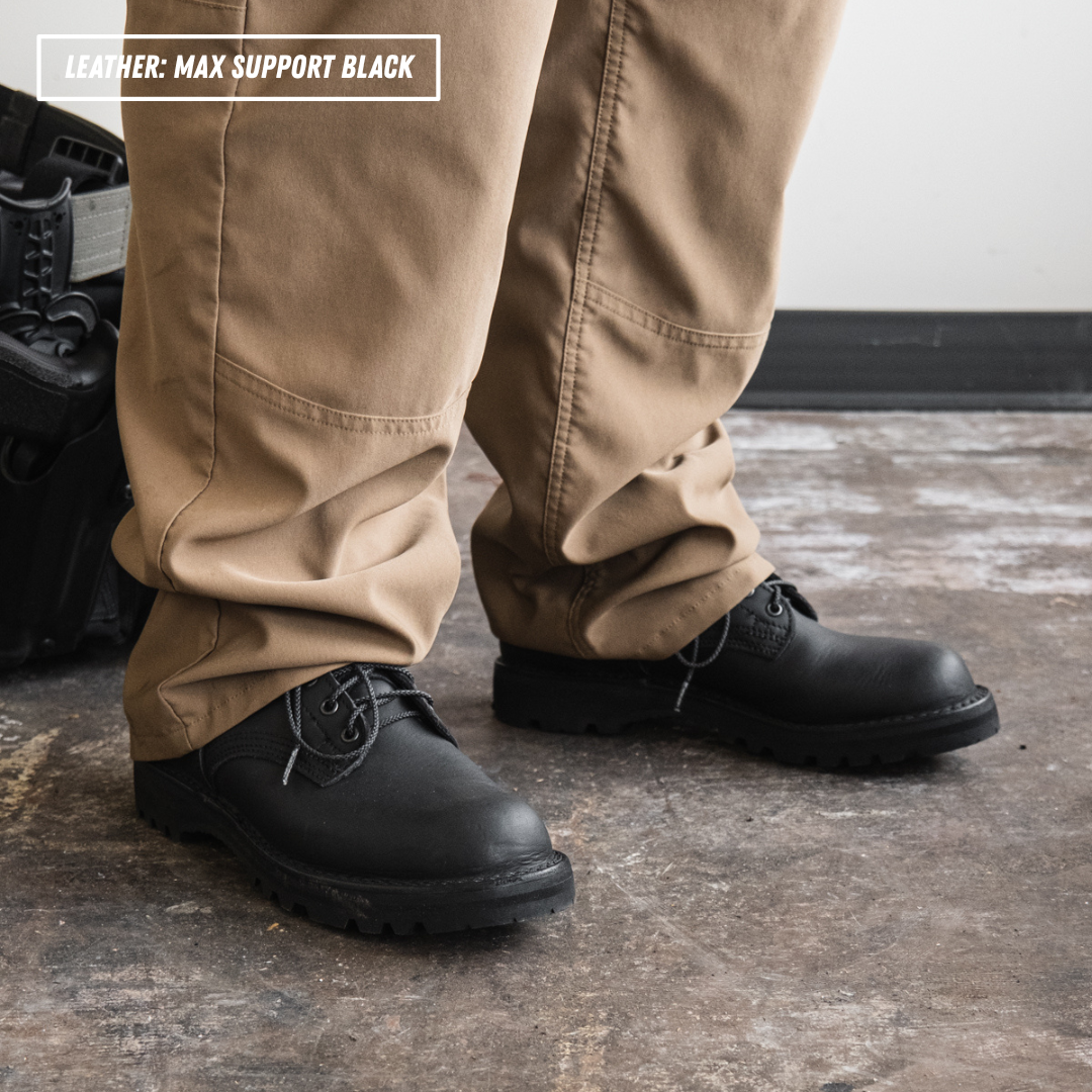 A close-up of a person's legs wearing tan work pants and black leather work boots on a concrete floor, with a text overlay in the top left that reads 'LEATHER - MAX SUPPORT BLACK'.