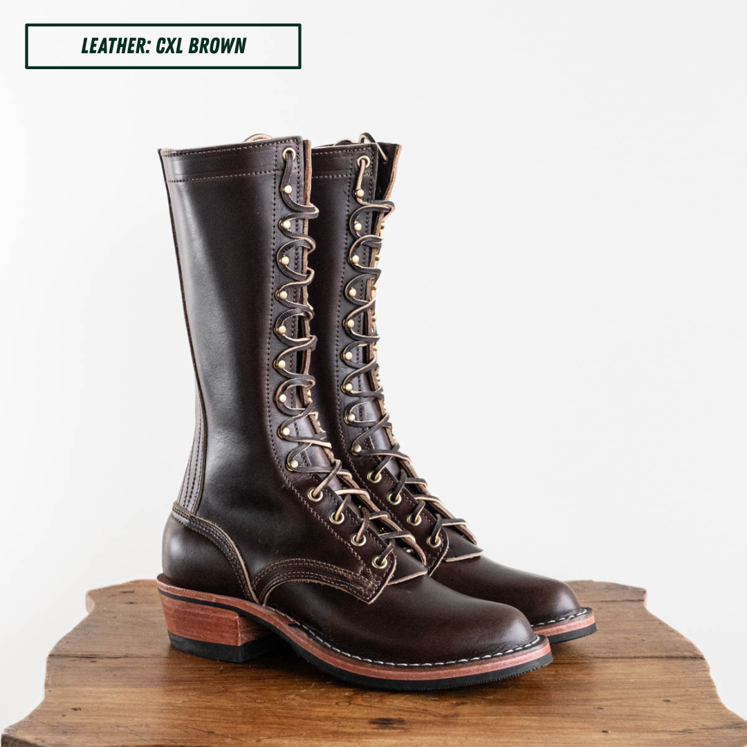 A pair of tall brown leather lace-up boots with metal eyelets and stacked wooden heels, displayed on a rustic wooden surface against a plain light background. A text label in the top left reads 'LEATHER: CXL BROWN'.
