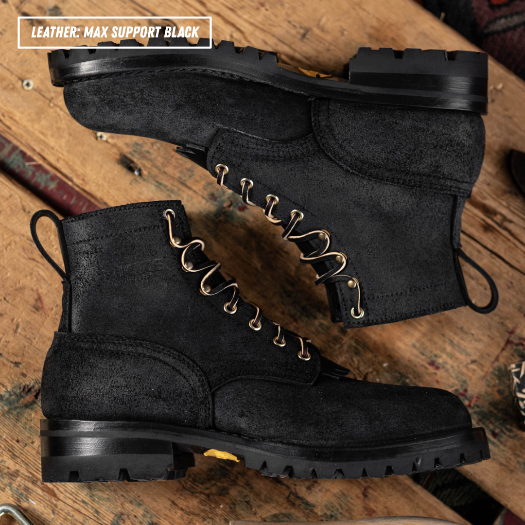 A pair of black leather lace-up boots with gold-tone metal eyelets, displayed on a rustic wooden surface. One boot is positioned above the other, showing the side profile and the top of the other boot.