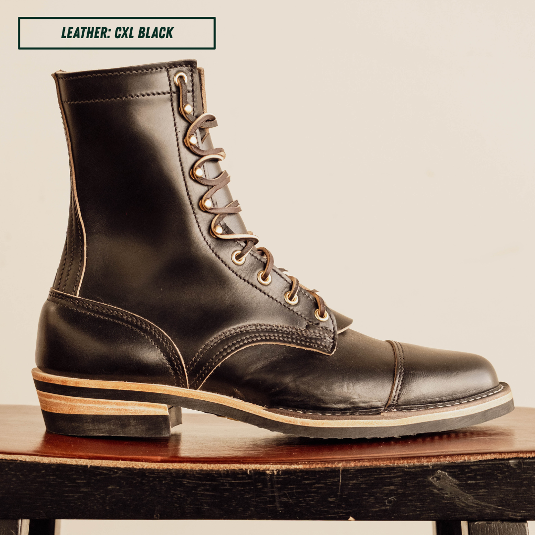 A single black leather lace-up ankle boot shown in side profile, resting on a wooden surface against a plain light beige background. A small label at the top reads 'LEATHER: CXL BLACK'.