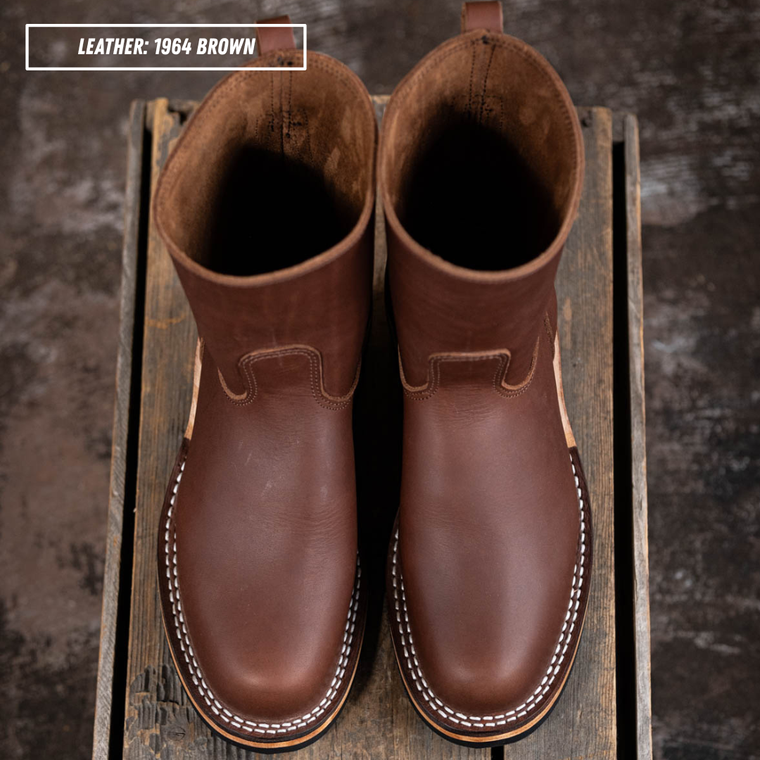 A pair of brown leather mid-calf boots with white contrast stitching, displayed on a rustic wooden crate. A text overlay in the top left corner reads 'LEATHER: 1964 BROWN'.