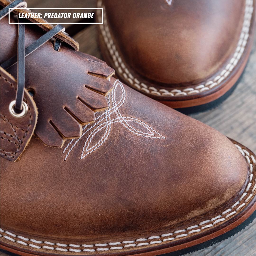 A close-up of a brown leather boot with white contrast stitching, fringe detail, and white embroidered stitching on the side. A text label in the top left reads 'LEATHER: PREDATOR ORANGE'.