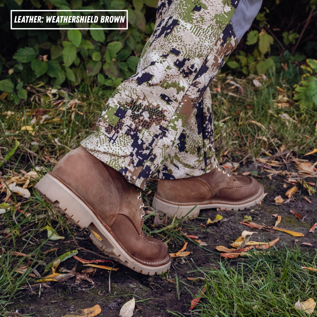 A person wearing camouflage pants and brown leather work boots standing outdoors on grass and dirt with scattered leaves. A text box in the top left reads 'LEATHER: WEATHERSHIELD BROWN'.
