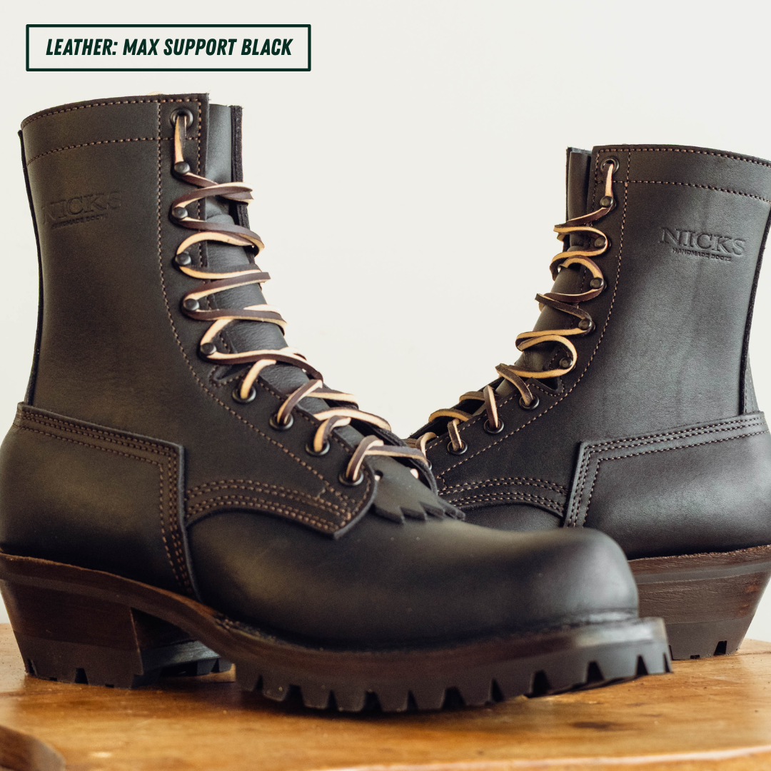 A pair of black leather lace-up work boots with metal eyelets and a rugged lug sole, displayed on a wooden surface against a light background. A text overlay in the top left reads 'LEATHER: MAX SUPPORT BLACK'.