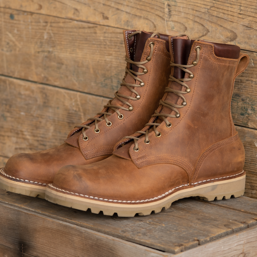A pair of brown leather lace-up work boots with off-white stitching and beige soles, displayed on a rustic wooden surface against a wooden plank background.