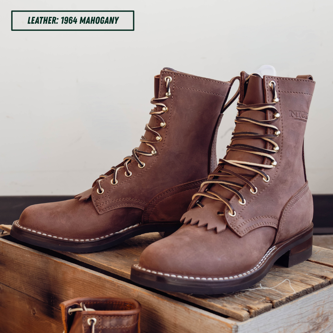 A pair of brown leather lace-up boots with fringe detailing and metal eyelets, displayed on a wooden crate against a plain wall. A text label in the top left reads 'LEATHER: 1964 MAHOGANY'.
