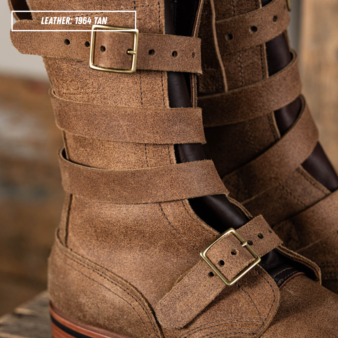 Close-up of a pair of tan leather double-strap boots with brass buckle details, shown on a rustic wooden surface. A text overlay in the top left reads 'LEATHER: 1964 TAN'.