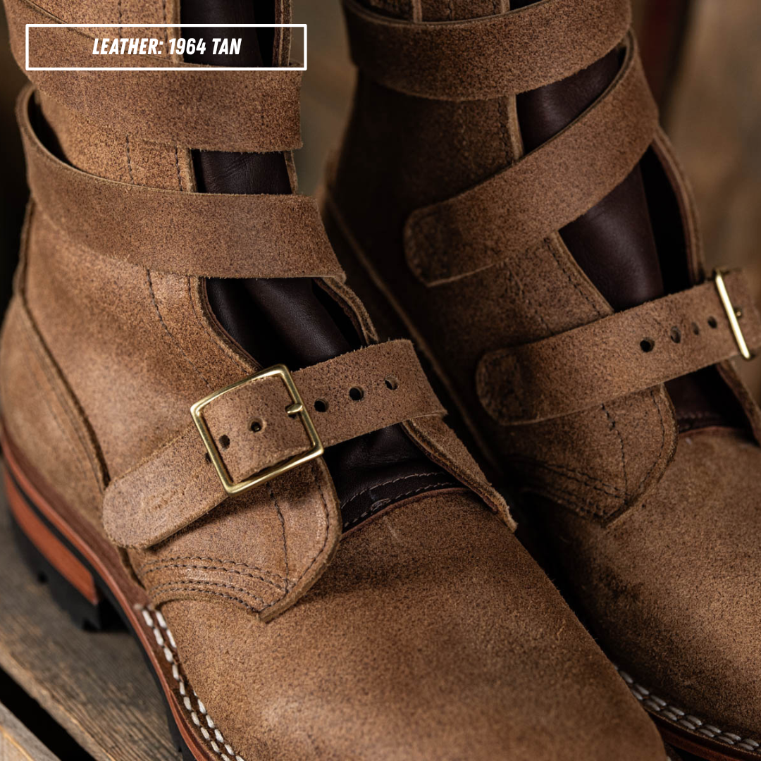 Close-up of a pair of tan leather double-strap boots with brass buckle details, shown on a rustic wooden surface. A text overlay in the top left reads 'LEATHER: 1964 TAN'.