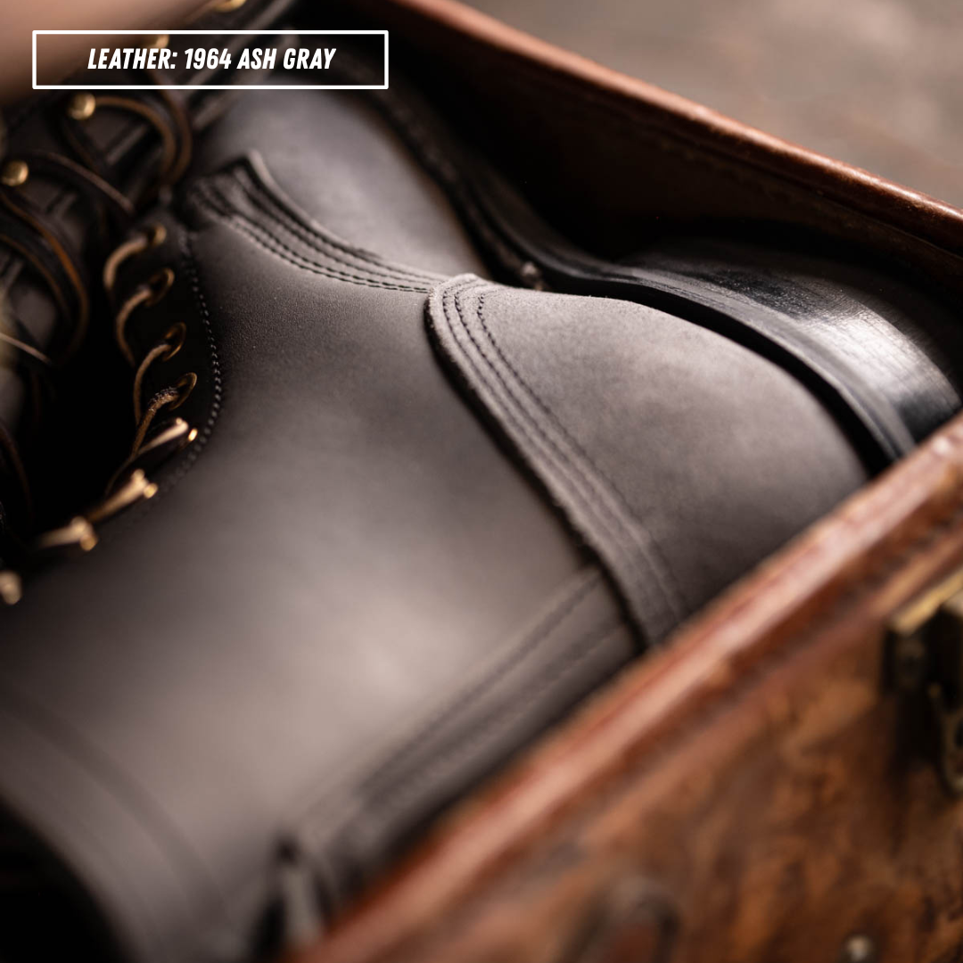 A close-up of a dark gray leather boot inside an open brown suitcase, with a text label in the top-left corner reading 'LEATHER: 1984 ASH GRAY'.