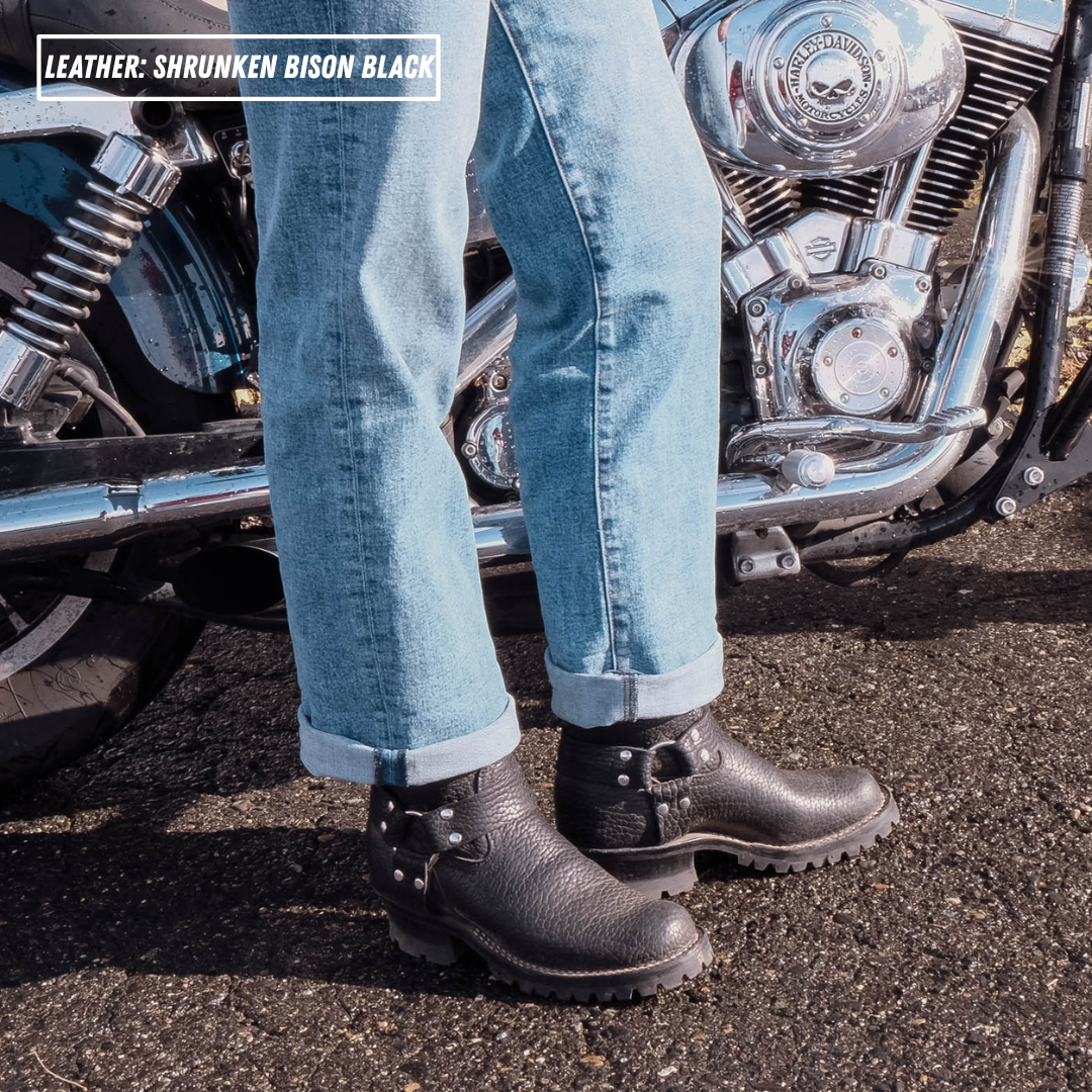 A person wearing cuffed light blue jeans and black leather motorcycle boots stands beside a chrome motorcycle on asphalt. A text overlay in the top left reads 'LEATHER: SHRUNKEN BISON BLACK'.
