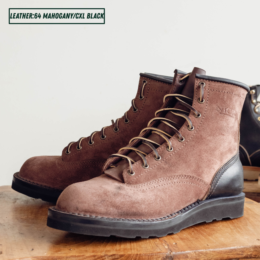 A pair of brown suede lace-up work boots with black leather single piece backstay sitting on wooden table. A text label in the top left reads 'LEATHER:64 MAHOGANY/CXL BLACK'.