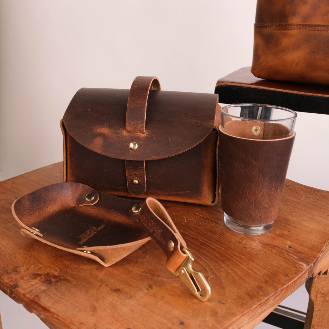 A dark brown leather organizer set on a rustic wooden table, including a small leather case with a top handle, a matching leather tray with a strap and brass clasp, a clear glass with a leather sleeve, and a partial leather bag in the background.