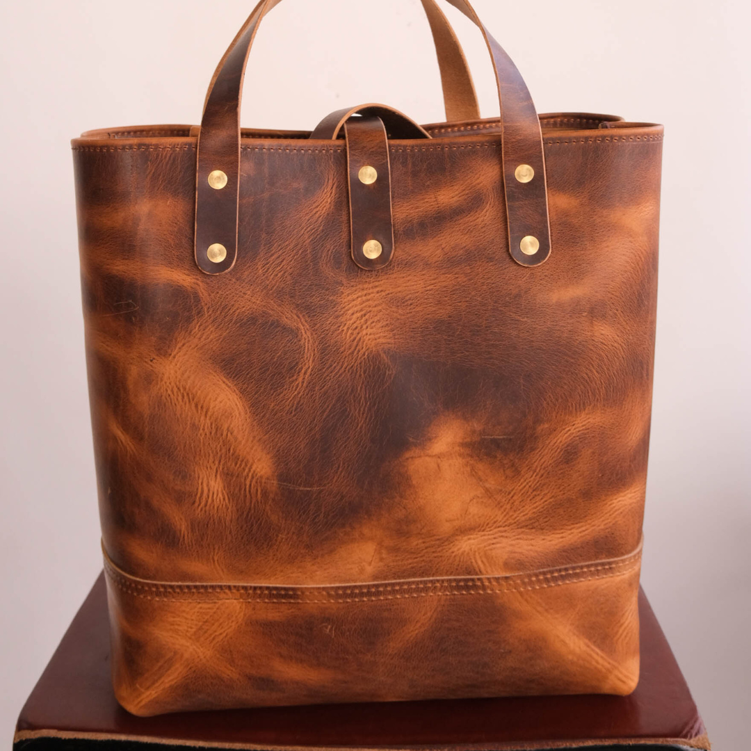 A large brown leather tote bag with distressed, marbled texture and brass-tone rivets, shown standing upright on a dark surface against a plain light background.