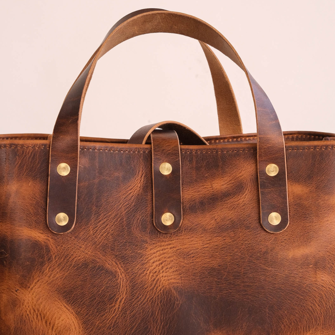 Close-up of a brown distressed leather tote bag with double top handles and brass rivets on a light beige background.