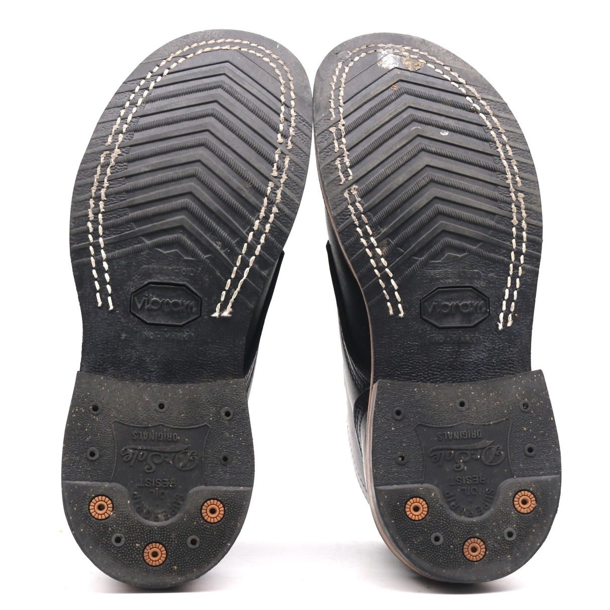 A pair of black leather boots shown from the sole side, featuring textured tread patterns, white contrast stitching, and orange lug nuts on a white background.