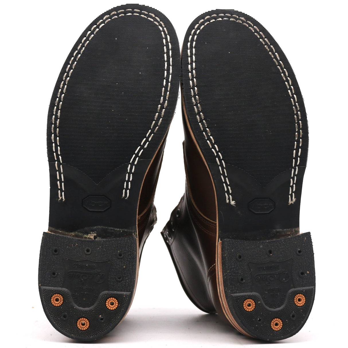 The soles of a pair of dark brown leather boots, showing the black tread, white contrast stitching, and orange rubber protective caps on the heels against a white background.