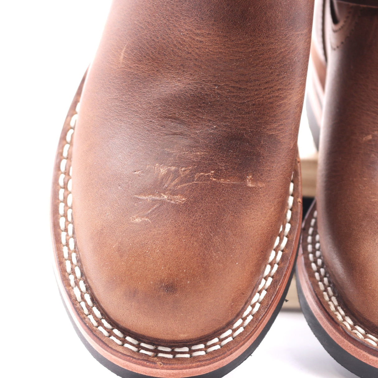 Close-up of a brown leather boot showing the vamp with visible scuffs and scratches on the leather surface, white contrast stitching around the edge, and part of the sole.