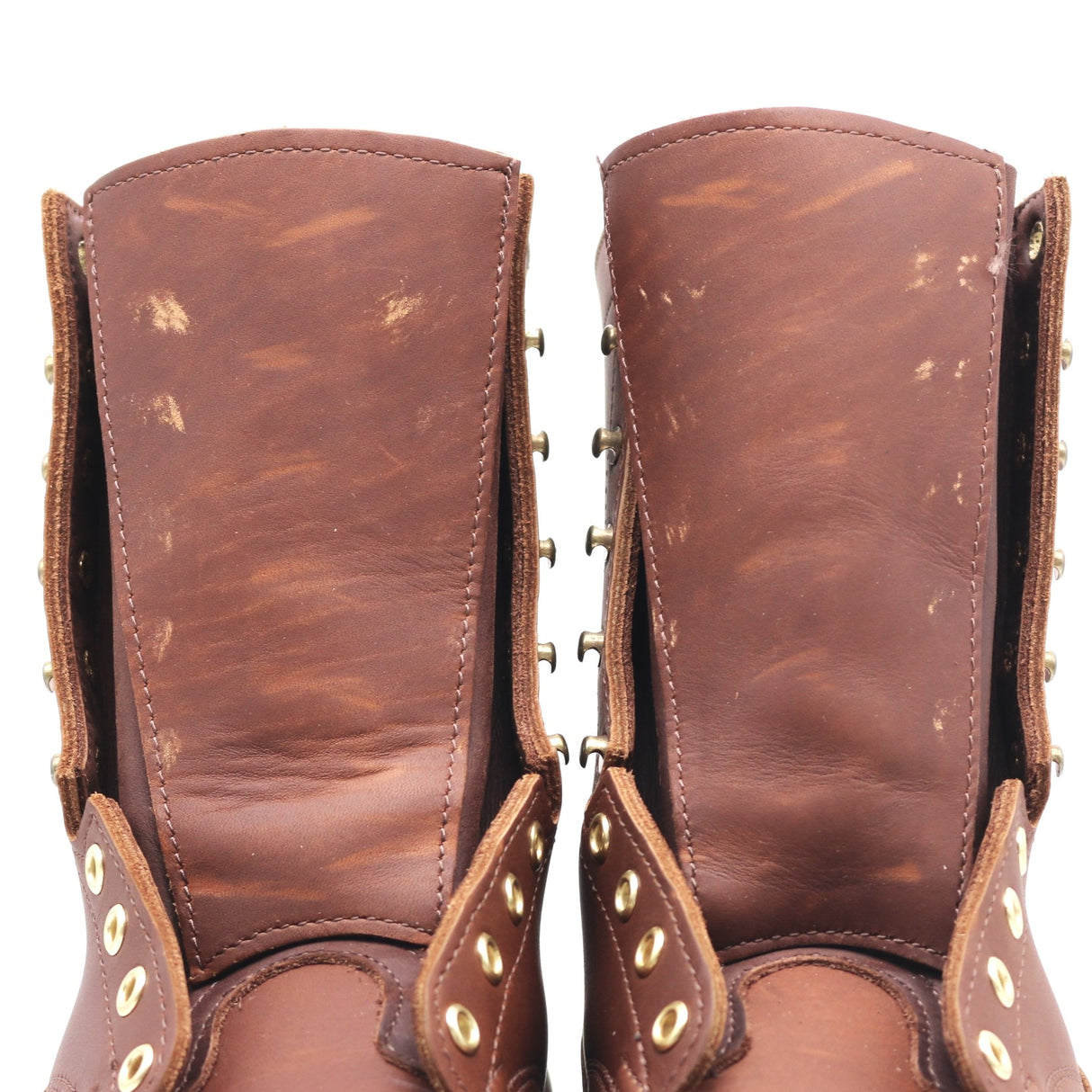 A close-up of a pair of distressed brown leather boots showing the inner shafts, worn leather texture, and brass eyelets and studs.