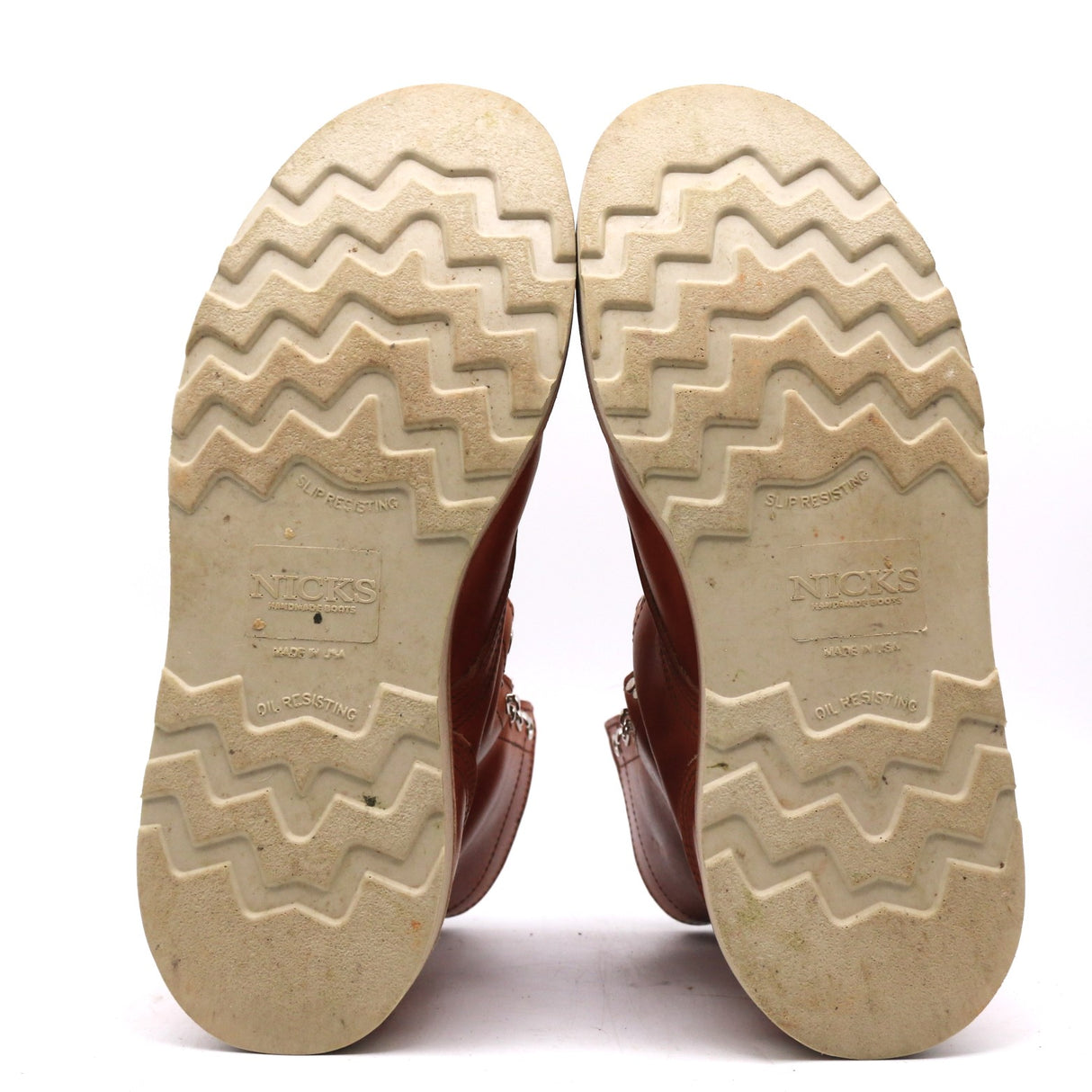 The soles of a pair of brown leather boots, shown from the bottom against a white background. The soles are a light beige/tan color with a zigzag tread pattern, and the brand name 'NICKS' is embossed in the center.