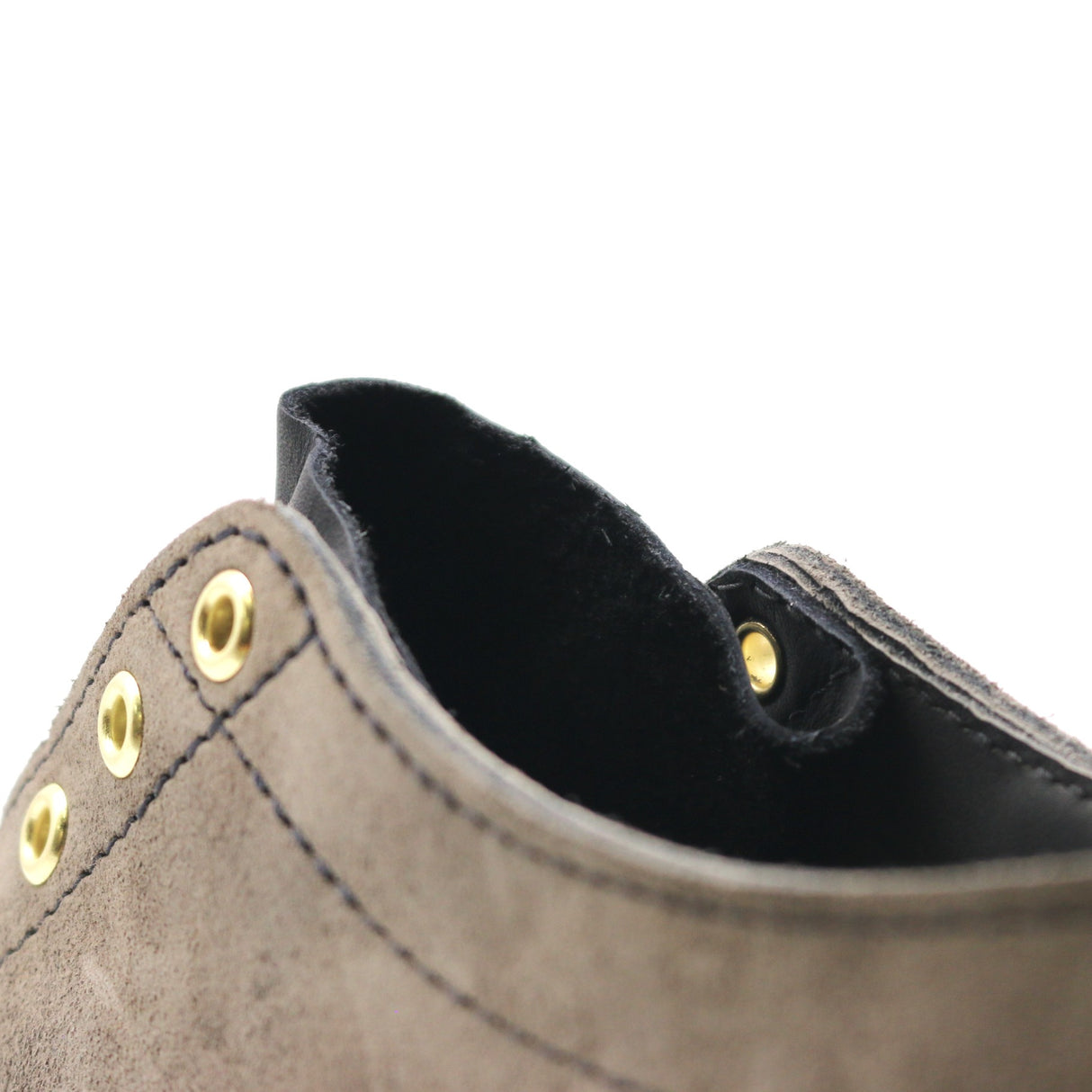 Close-up detail of a taupe suede shoe with gold-tone metal grommets and a black interior lining, set against a plain white background.