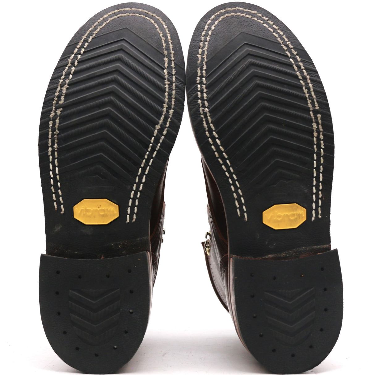 The soles of a pair of dark brown leather boots, showing the black treaded rubber outsoles with white contrast stitching and yellow Vibram logos.