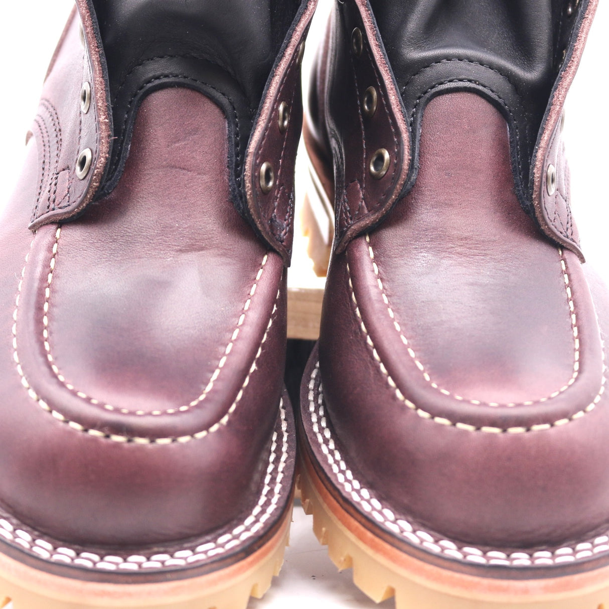 Close-up of a pair of dark burgundy leather boots with white contrast stitching and metal eyelets, shown against a white background.