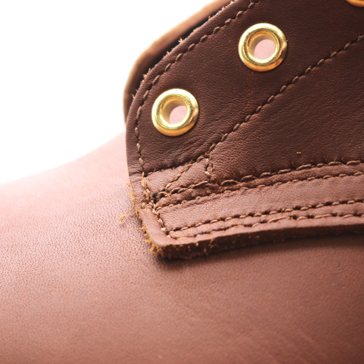 Close-up of a brown leather boot or shoe showing the texture of the leather, a stitched tab, and two brass grommets on a white background.