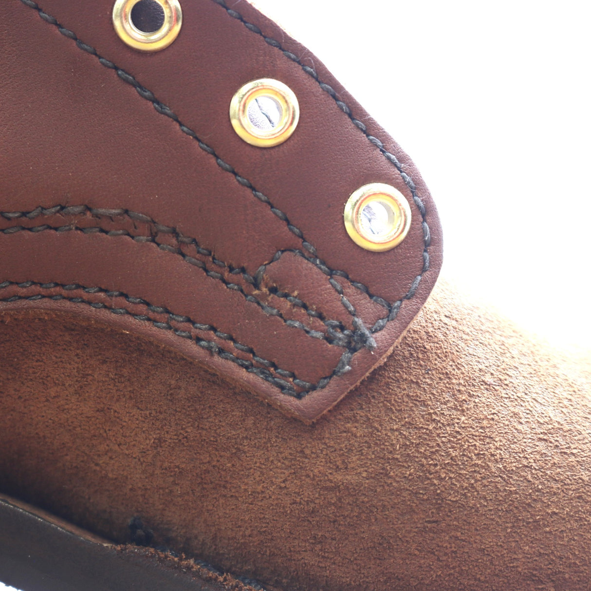 Close-up detail of a brown leather shoe or boot showing a stitched overlay panel with brass grommets and a suede-like brown footbed.