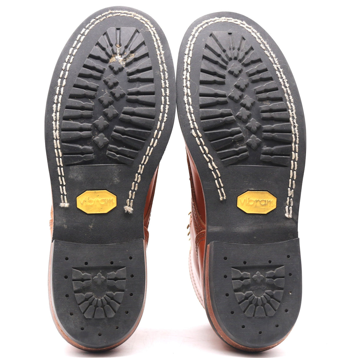 A pair of brown leather boots shown from the sole, displaying the black rubber tread with white stitching and yellow Vibram logos on a white background.