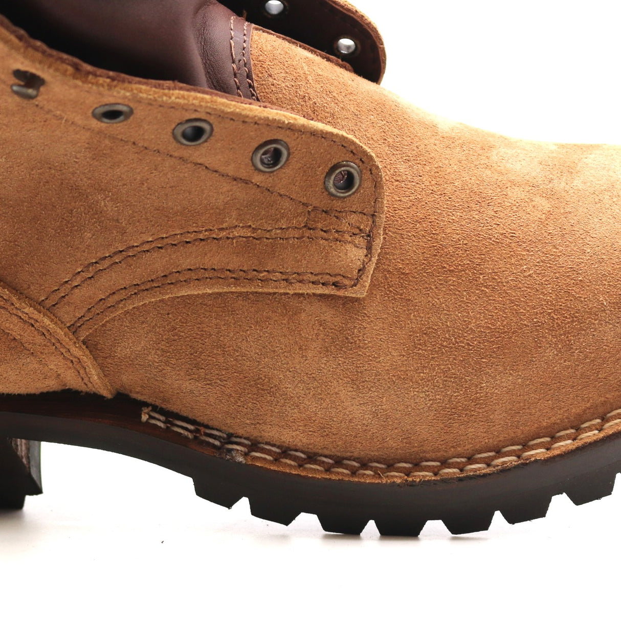 A close-up side view of a tan suede work boot with dark brown stitching, metal eyelets, and a black lug sole on a white background.