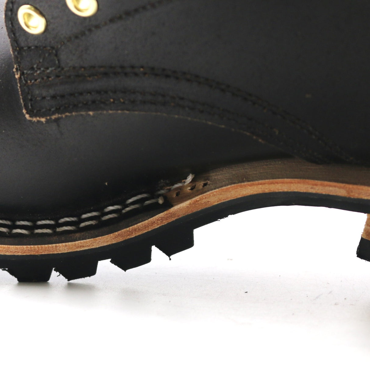 Close-up of a black leather lace-up boot showing the side profile, including brass eyelets, stitching, a tan midsole, and a black lug sole on a white background.
