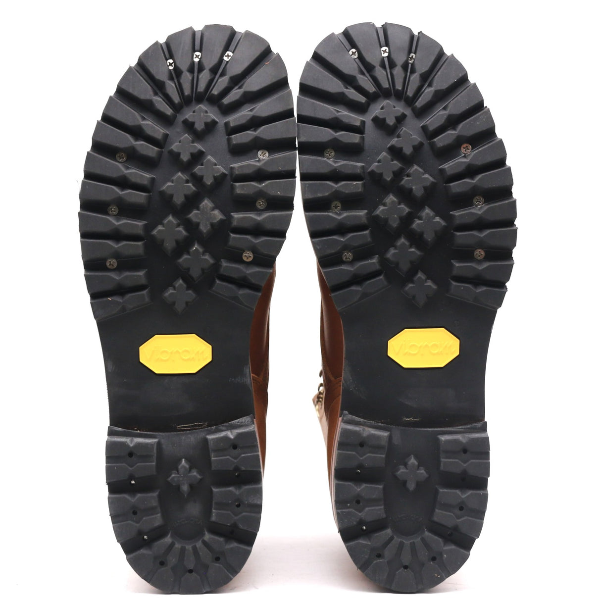 A pair of brown boots shown from the bottom, displaying their black Vibram soles with yellow Vibram logos and metal spikes.
