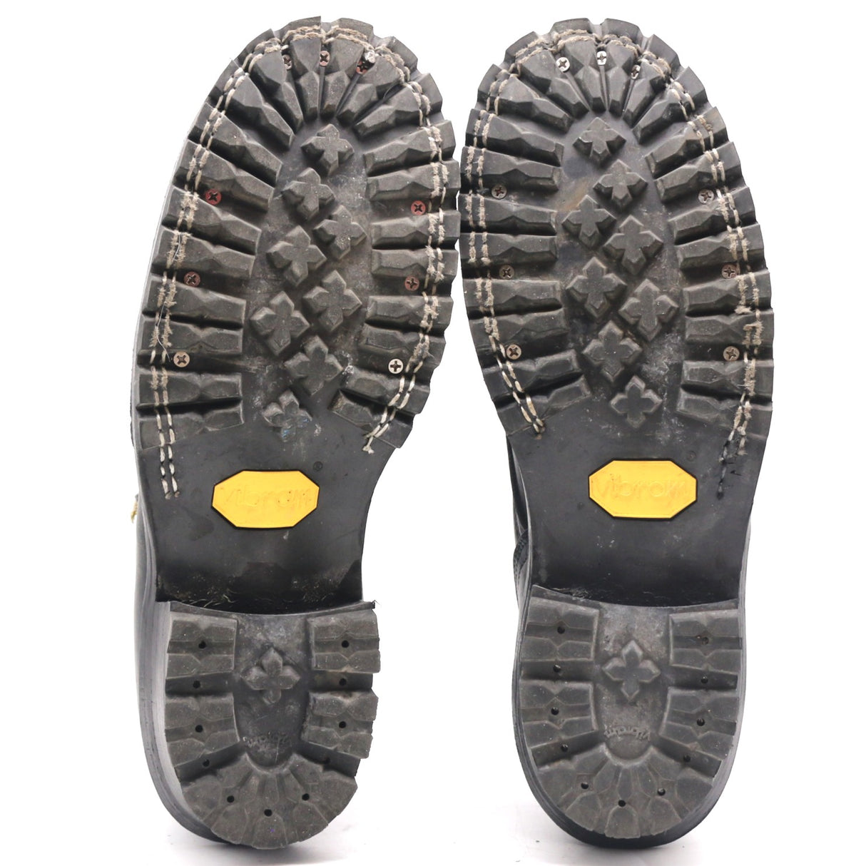 A pair of black heavy-duty boots shown from the sole, displaying deep lugs, stitching, and yellow Vibram logos on a white background.