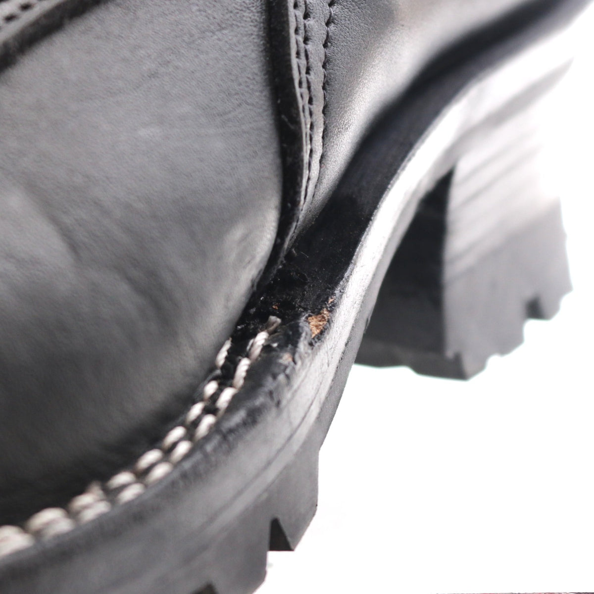 Close-up of a black leather boot corner showing a worn, scuffed edge between the leather vamp and the black lug sole, with white stitching along the edge.