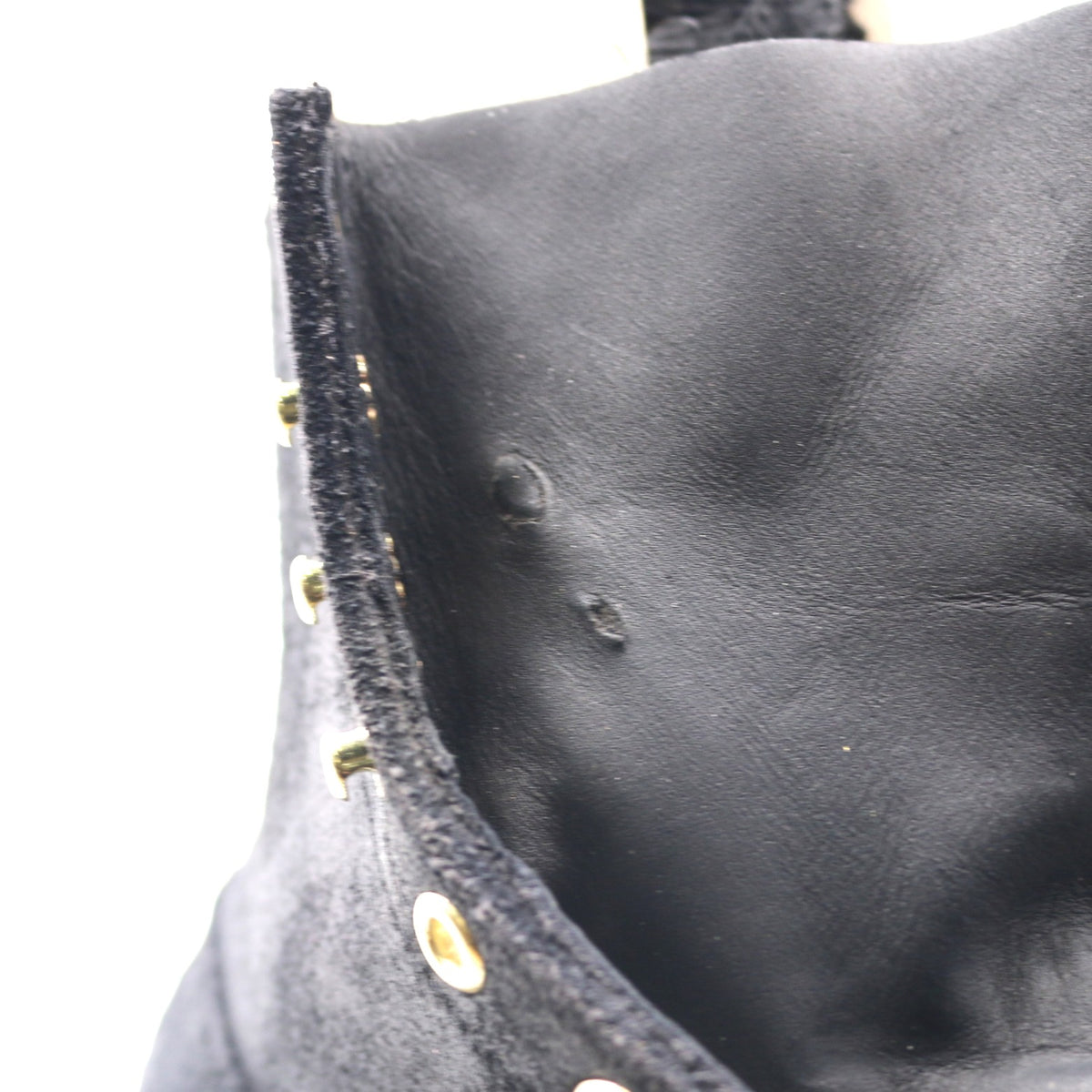 Close-up detail of a black leather garment, showing the texture of the leather, a black velvet trim with gold-tone metal studs, and a white background.