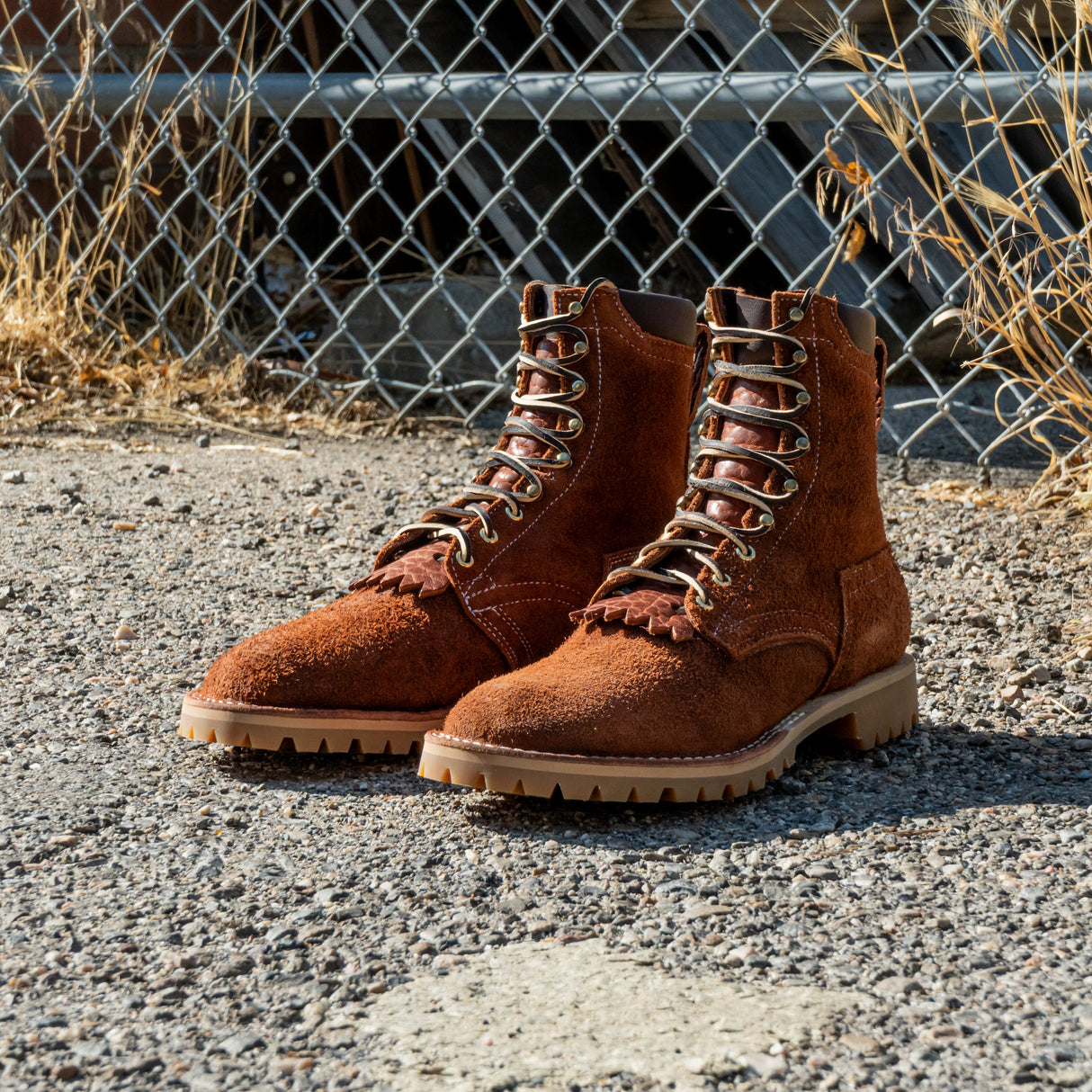 Shrunken bison brown leather Bear Slayer with brown padded collar, honey vibram outsole and 4 relief notches. Boots sitting on gravel outside in front of chain link fence.