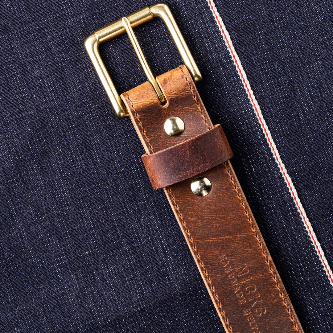 A close-up of a brown leather watch strap with a brass buckle, laid diagonally on dark navy denim fabric with red and white contrast stitching.