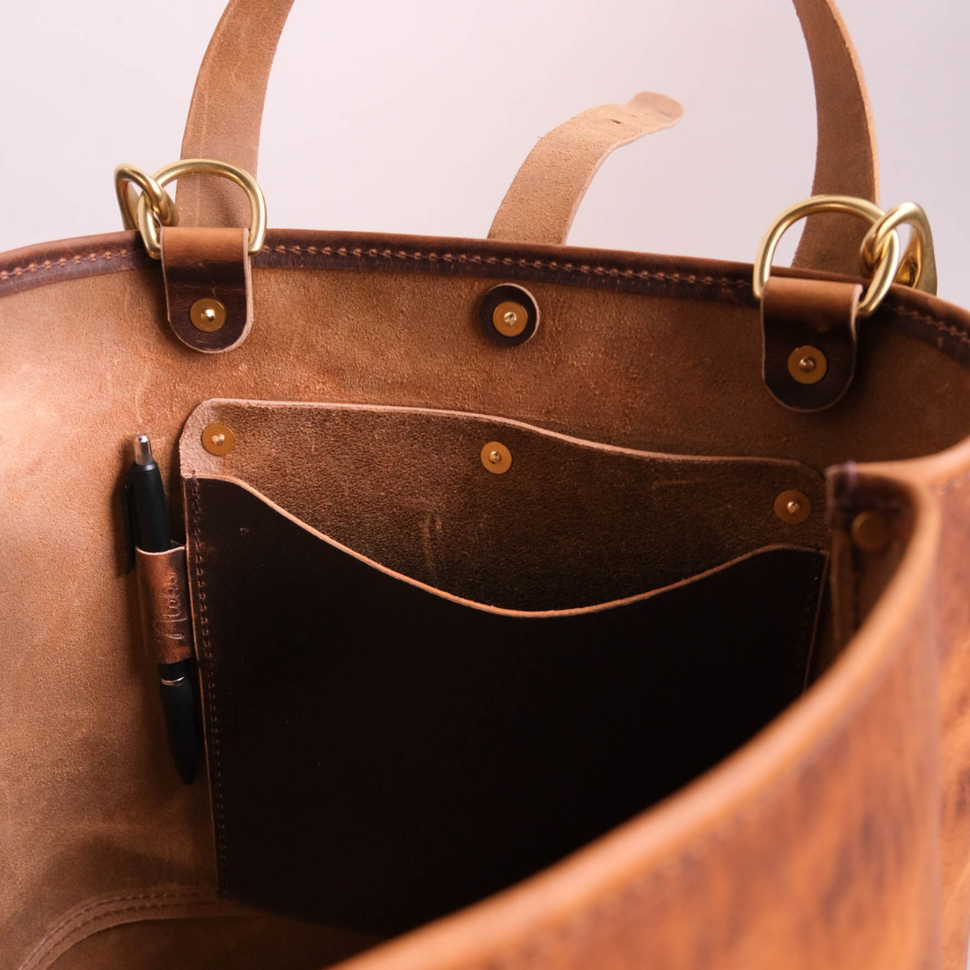Close-up of the interior of a brown leather tote bag, showing a pen in a side pocket and a dark insert pocket, with brass-tone hardware and handles against a light neutral background.