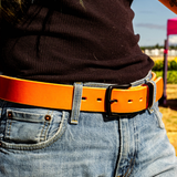 A close-up of a person wearing a bright orange leather belt with a black buckle, paired with blue jeans and a black ribbed top, outdoors.