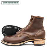 A pair of brown leather lace-up ankle boots shown on a white background, with one boot standing upright and the other lying on its side to display the black outsole. A text label in the top left reads 'LEATHER: SHACKLETON COGNAC'.