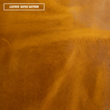 A close-up texture shot of smooth leather in the color 'Bayou Saffron', showing a warm golden-brown hue with subtle natural grain and folds. A white label in the top-left corner reads 'LEATHER: BAYOU SAFFRON'.